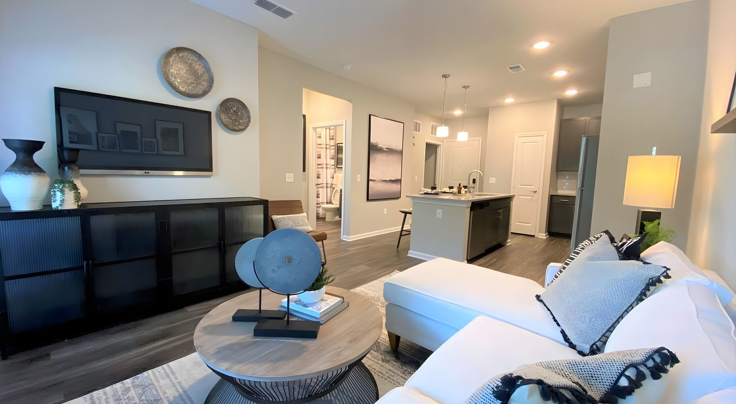 Modern living room with a white sectional sofa, decorative pillows, a round coffee table, and an open kitchen in the background.