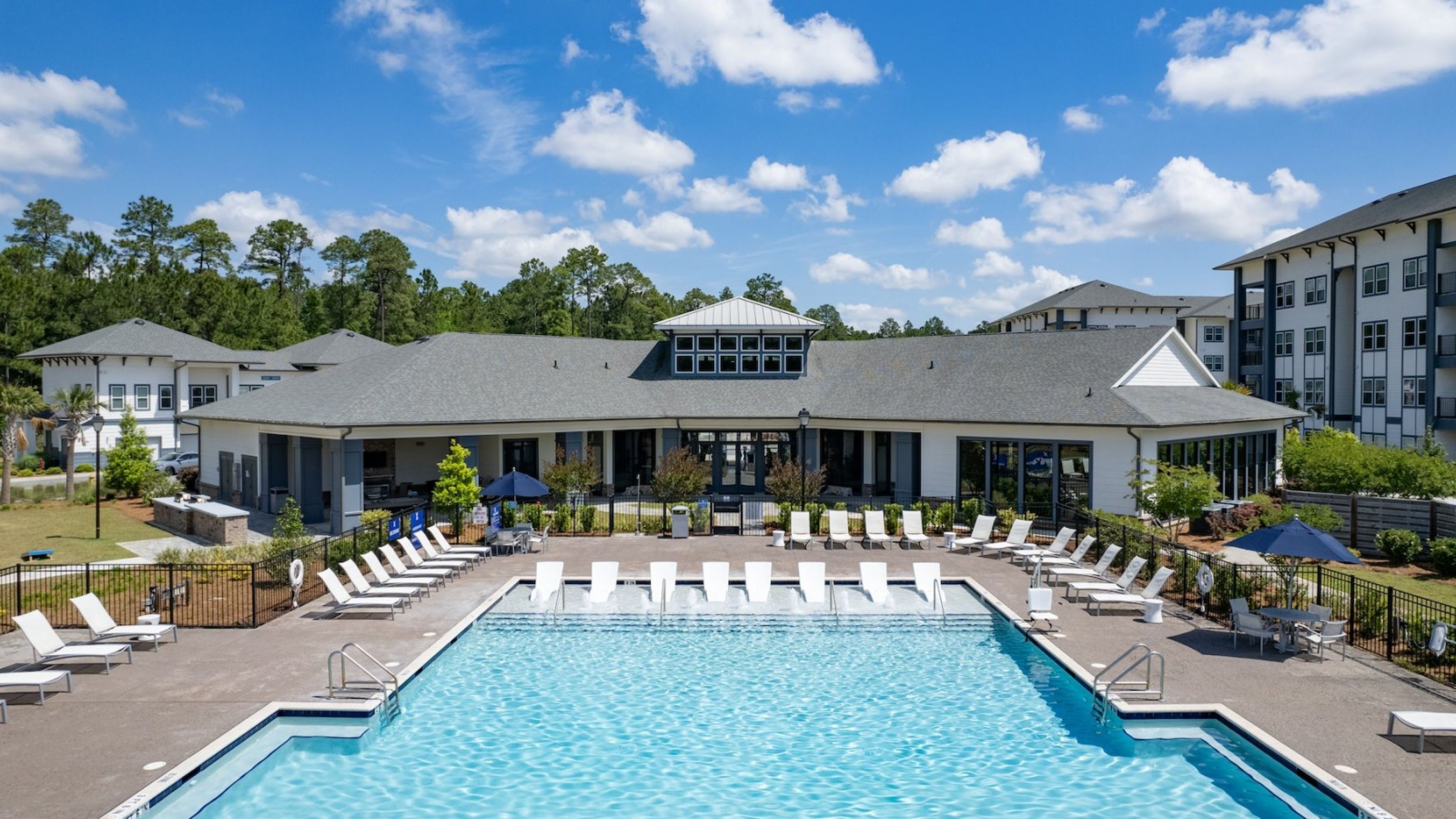 A bright, sunny pool area featuring a large swimming pool, lounge chairs, and a modern clubhouse surrounded by lush greenery.