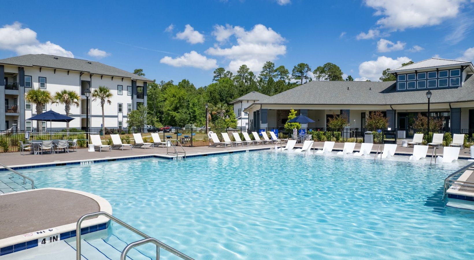 A bright outdoor swimming pool surrounded by lounge chairs, palm trees, and a modern apartment building under a clear blue sky.