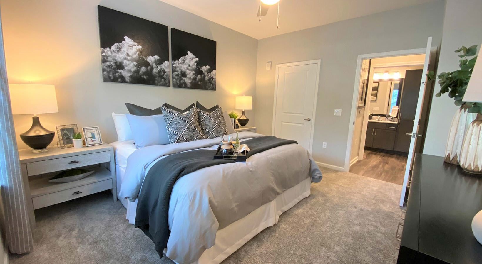 A cozy bedroom featuring a large bed with decorative pillows, a bedside table with a lamp, and access to a bathroom. Soft gray tones dominate.